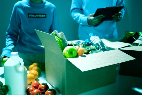 Deux personnes devant un carton rempli de courses, avec des légumes, une bouteille d’eau et des boîtes de conserve, symbolisant le service d’aide à la vie quotidienne d’ADATTA.